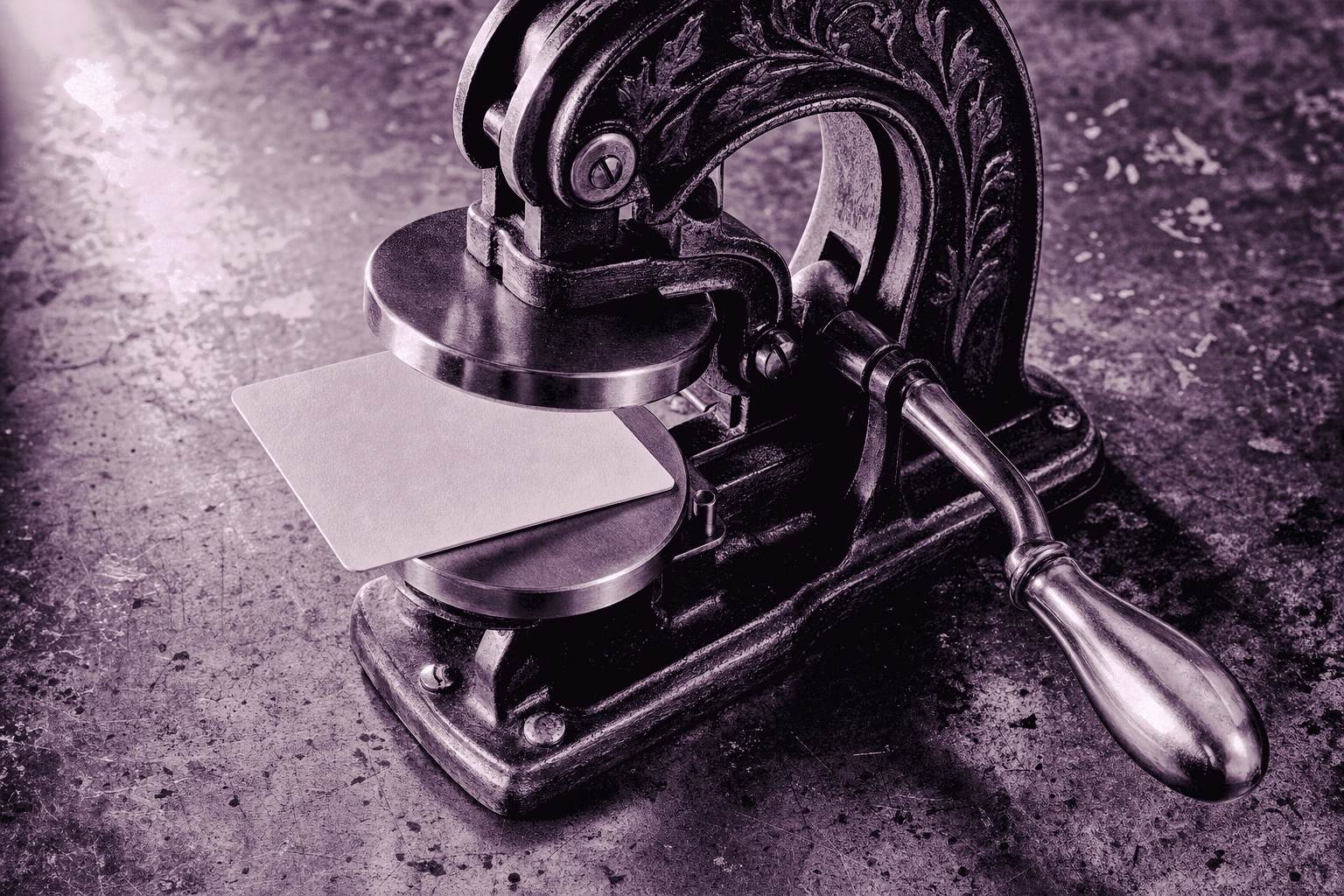 A blank unembossed card blank resting in the open jaws of a hand-cranked embossing press on a scarred steel bench